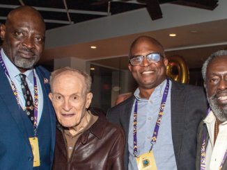 Four men pose together at an indoor event, wearing lanyards and smiling at the camera.