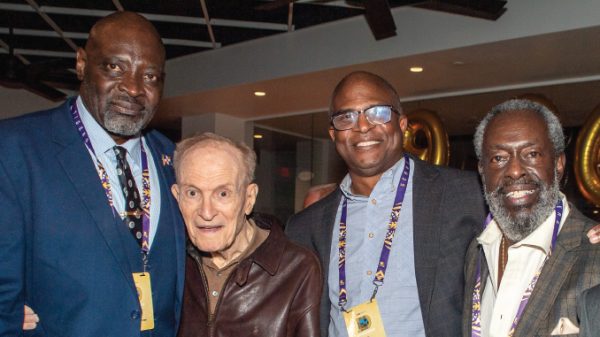 Four men pose together at an indoor event, wearing lanyards and smiling at the camera.