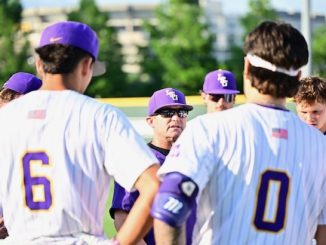 Baseball team in white uniforms with purple caps huddles on a sunny field, listening to a coach.