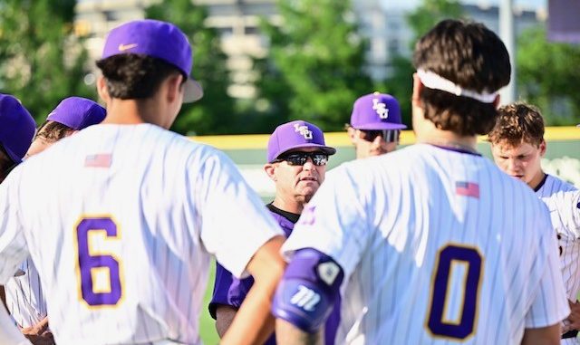Baseball team in white uniforms with purple caps huddles on a sunny field, listening to a coach.