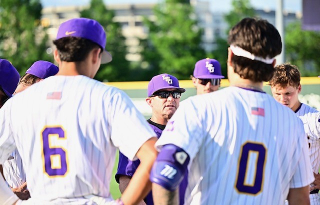 Baseball team in white uniforms with purple caps huddles on a sunny field, listening to a coach.