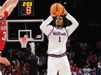 Basketball player in a white Wildcats jersey with #1 shoots a high-arc jumper during a game; crowd and scoreboard visible in the background.