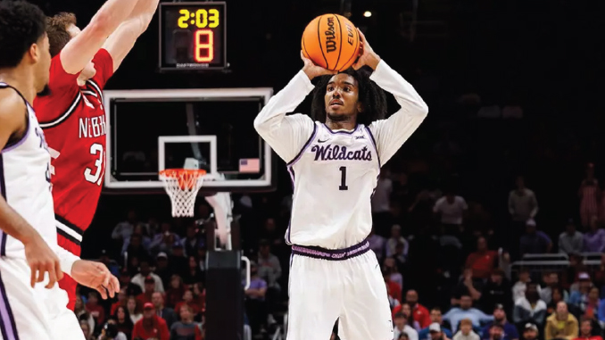 Basketball player in a white Wildcats jersey with #1 shoots a high-arc jumper during a game; crowd and scoreboard visible in the background.
