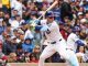 Cubs batter in pinstriped uniform swings at a pitch during a game as a catcher and cheering crowd look on behind him.