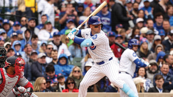 Cubs batter in pinstriped uniform swings at a pitch during a game as a catcher and cheering crowd look on behind him.