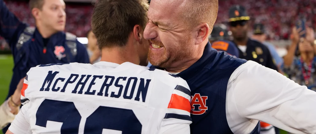 Two football players embrace in a celebratory hug on the field, one wearing a white and navy jersey with 'McPherson' on the back and the other in blue.