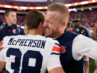 Two football players embrace in a celebratory hug on the field, one wearing a white and navy jersey with 'McPherson' on the back and the other in blue.