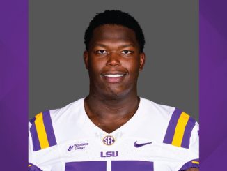 Portrait of an LSU football player wearing a white jersey with purple and gold stripes, against a gray backdrop