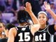 Female basketball players in black jerseys high-fiving during a game, with the number 15 visible on one player's back and a purple-clad crowd in the background.