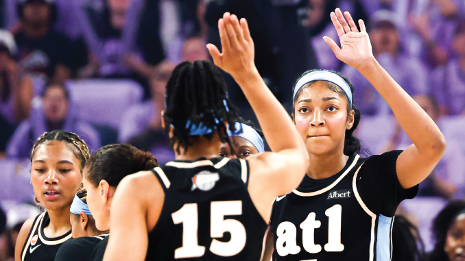 Female basketball players in black jerseys high-fiving during a game, with the number 15 visible on one player's back and a purple-clad crowd in the background.