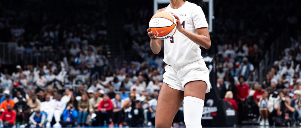 Female basketball player in white uniform holding an orange basketball, looking to her left on a crowded indoor court.
