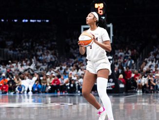 Female basketball player in white uniform holding an orange basketball, looking to her left on a crowded indoor court.