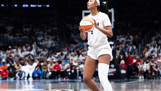 Female basketball player in white uniform holding an orange basketball, looking to her left on a crowded indoor court.