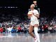 Female basketball player in white uniform holding an orange basketball, looking to her left on a crowded indoor court.