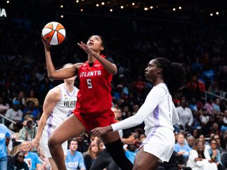 Basketball players in red and white uniforms jump for the ball amid a crowded arena; focus on Atlanta player reaching for a rebound.