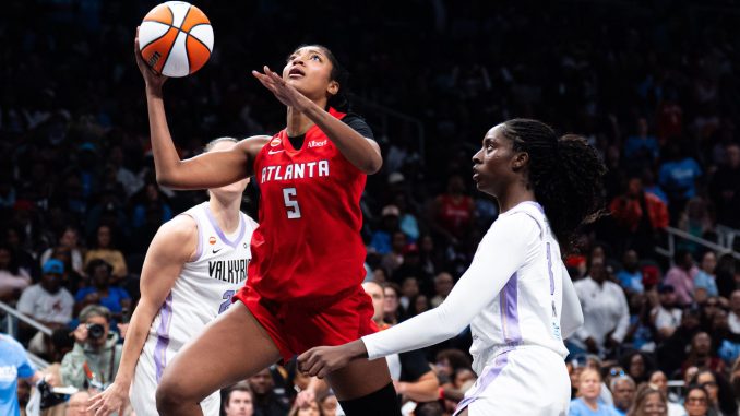 Basketball players in red and white uniforms jump for the ball amid a crowded arena; focus on Atlanta player reaching for a rebound.