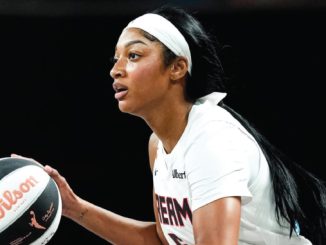 Female basketball player in a white uniform with a headband, holding a basketball and looking ahead.