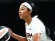 Female basketball player in a white uniform with a headband, holding a basketball and looking ahead.