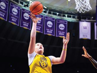 Basketball player in a yellow Tigers jersey leaps for a shot near the basket, NCAA banners visible in the arena.