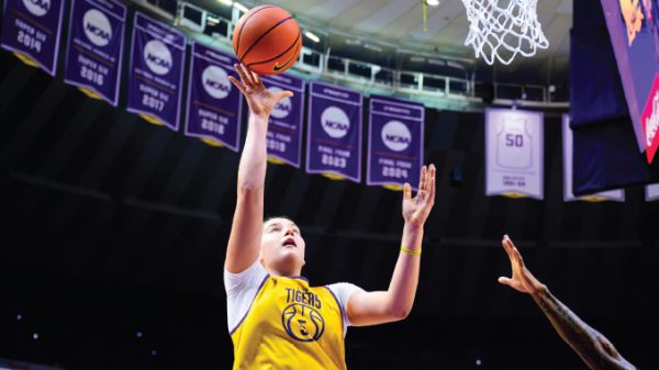 Basketball player in a yellow Tigers jersey leaps for a shot near the basket, NCAA banners visible in the arena.