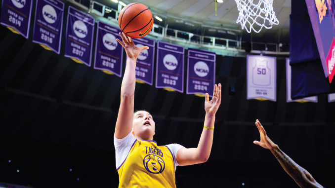 Basketball player in a yellow Tigers jersey leaps for a shot near the basket, NCAA banners visible in the arena.