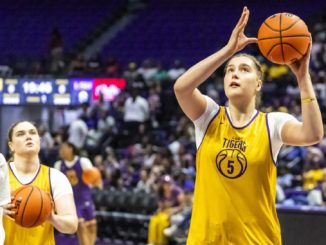 LSU Tigers women’s basketball players in yellow jerseys on the court; foreground player shoots a jump shot as teammates practice nearby in a gym.
