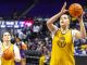LSU Tigers women’s basketball players in yellow jerseys on the court; foreground player shoots a jump shot as teammates practice nearby in a gym.
