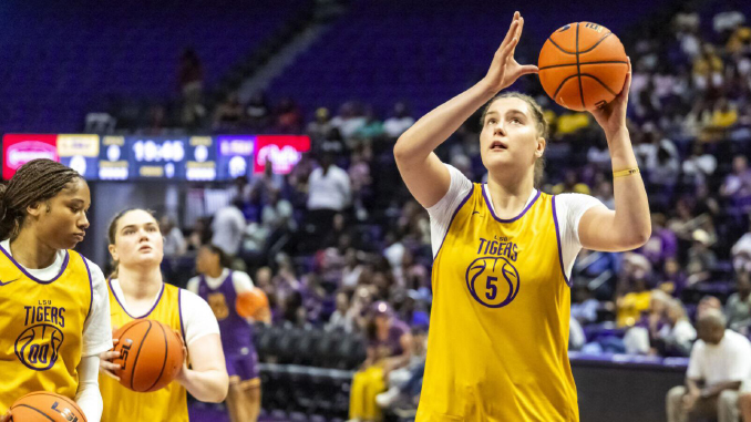 LSU Tigers women’s basketball players in yellow jerseys on the court; foreground player shoots a jump shot as teammates practice nearby in a gym.