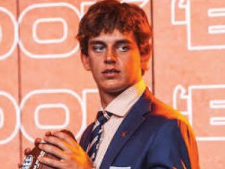 Young man in a blue suit and striped tie on a stage with orange neon signage in the background, holding a bottle.