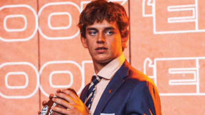Young man in a blue suit and striped tie on a stage with orange neon signage in the background, holding a bottle.