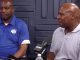 Two men sit at a podcast table with microphones; one in a blue polo, the other in a light gray polo, speaking to the camera side by side.