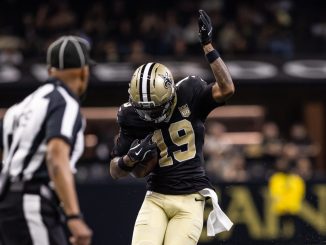 New Orleans Saints player in black and gold celebrates with his right arm raised on the field, an official nearby in view.