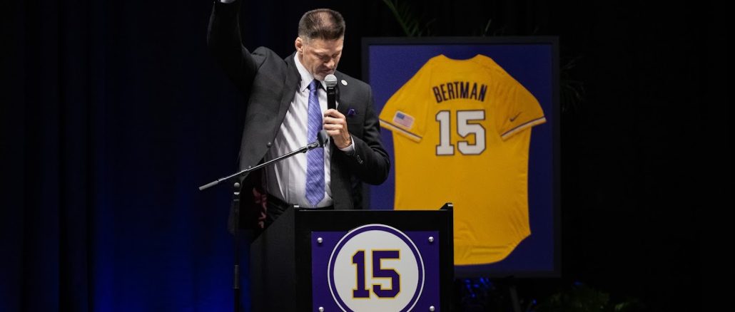 Man in a suit speaks at a podium, raising one hand while holding a mic, with a framed yellow jersey #15 behind him on stage.