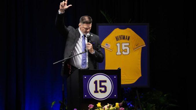 Man in a suit speaks at a podium, raising one hand while holding a mic, with a framed yellow jersey #15 behind him on stage.