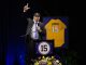 Man in a suit speaks at a podium, raising one hand while holding a mic, with a framed yellow jersey #15 behind him on stage.