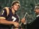 Young football player in a purple and gold jersey shakes hands with a suited man as he receives a trophy at an award ceremony.