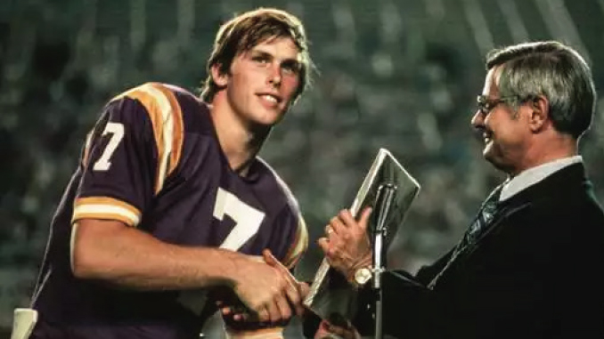 Young football player in a purple and gold jersey shakes hands with a suited man as he receives a trophy at an award ceremony.