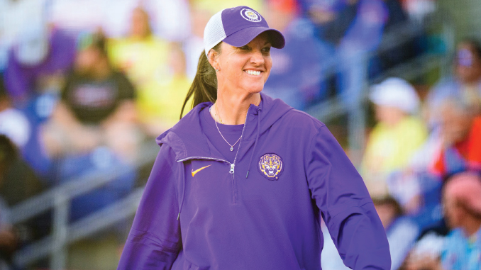 Smiling woman in a purple jacket and cap, standing at an outdoor sporting event with blurred spectators in the background.
