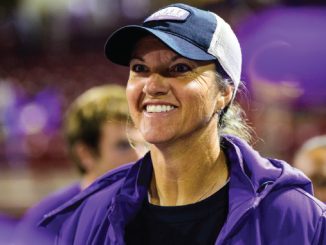 Smiling woman wearing a purple jacket and cap at an indoor event; purple banners blurred in the background.
