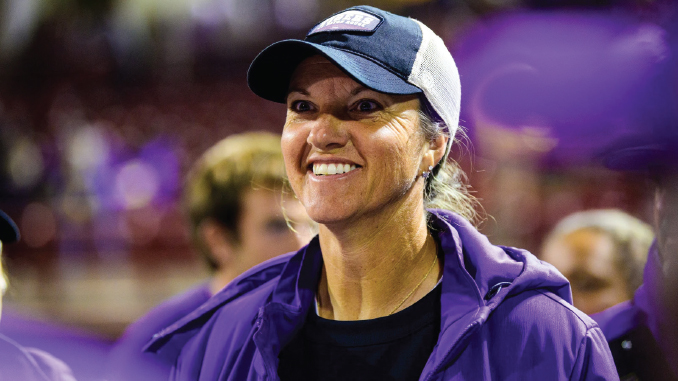 Smiling woman wearing a purple jacket and cap at an indoor event; purple banners blurred in the background.