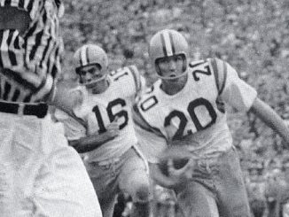Black-and-white action shot of two football players in white jerseys with numbers 15 and 20 chasing the ball as a referee watches nearby.
