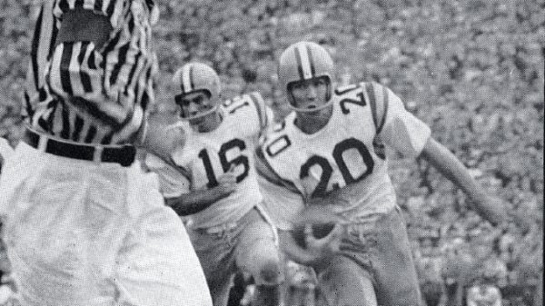 Black-and-white action shot of two football players in white jerseys with numbers 15 and 20 chasing the ball as a referee watches nearby.