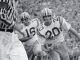 Black-and-white action shot of two football players in white jerseys with numbers 15 and 20 chasing the ball as a referee watches nearby.
