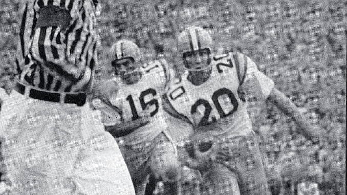 Black-and-white action shot of two football players in white jerseys with numbers 15 and 20 chasing the ball as a referee watches nearby.