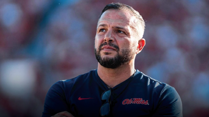 Man with a beard wearing an Ole Miss navy polo, sunglasses hanging from the collar, outdoors with a blurred stadium crowd behind him.