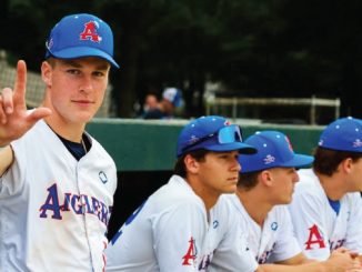 Baseball players in white uniforms sit on a dugout bench; foreground player faces camera and makes an L-shaped gesture with his hand toward the viewer.