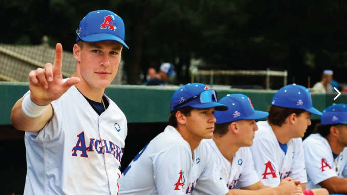 Baseball players in white uniforms sit on a dugout bench; foreground player faces camera and makes an L-shaped gesture with his hand toward the viewer.