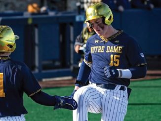 Two Notre Dame baseball players in blue and gold helmets high-five on the field, one wearing No. 15 and the other No. 11.