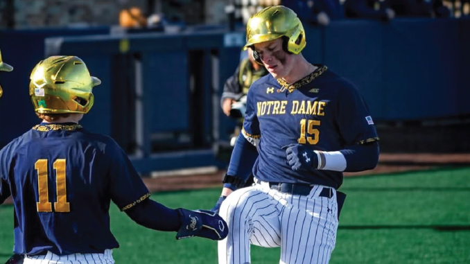 Two Notre Dame baseball players in blue and gold helmets high-five on the field, one wearing No. 15 and the other No. 11.