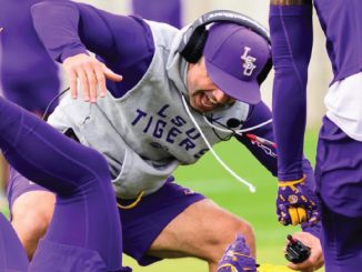 LSU Tigers football player in a purple cap and gray sweatshirt bends low during a training drill on the field.